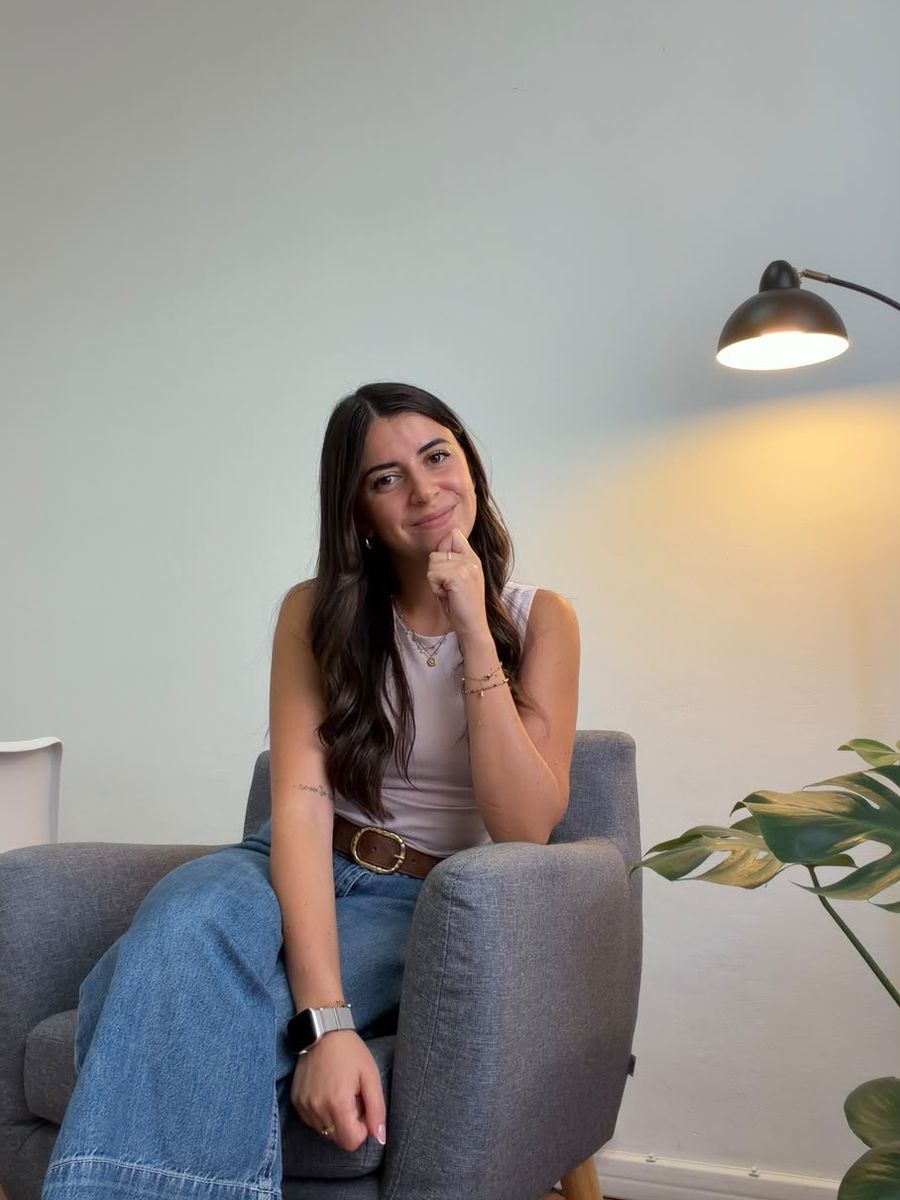 Sabrina Pili seduta su una poltrona grigia nel suo studio, accanto a una lampada accesa e a una pianta di monstera