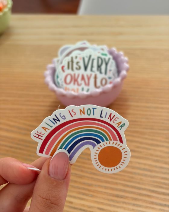 Una mano che tiene un adesivo con un arcobaleno e la scritta «Healing is not linear», sopra una ciotola di adesivi