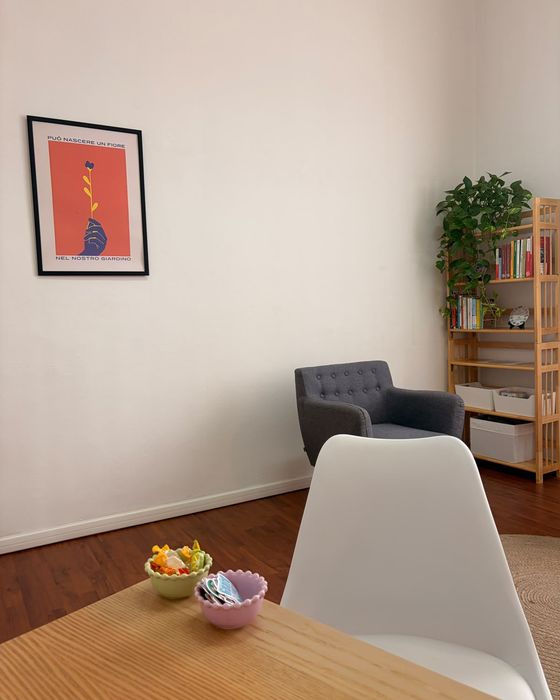 Un'altra parete dello studio: il poster «Può nascere un fiore nel nostro giardino», una poltrona grigia, una libreria con una pianta e un tavolo